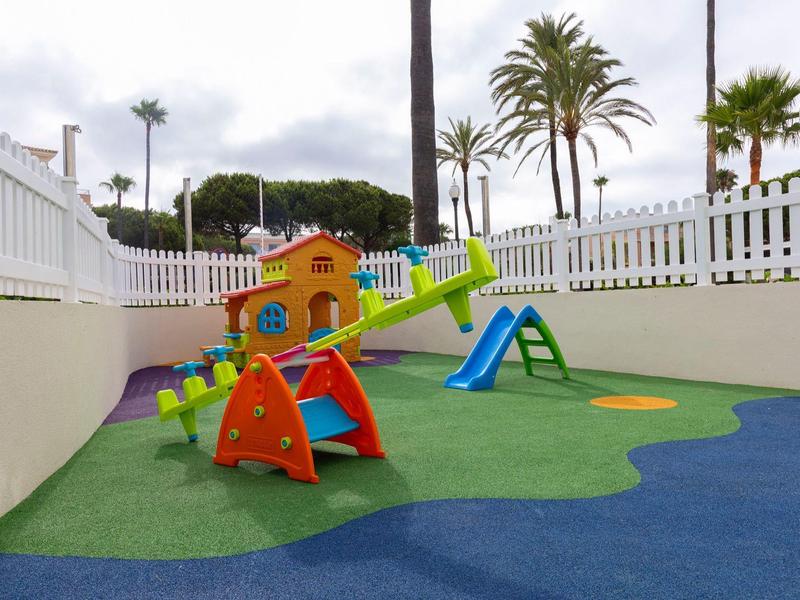 Kinderspielplatz mit bunten Spielgeräten, Rutsche, Klettergerüst und Umzäunung bei bewölktem Himmel.