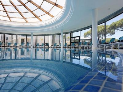 Innenaufnahme eines runden Hallenbads mit Glasdach und großen Fenstern, umgeben von Liegestühlen.
