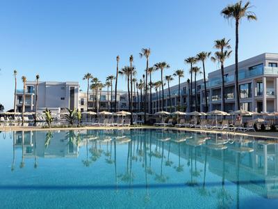 Moderner Hotelkomplex mit großem Pool und Palmen unter klarem blauem Himmel.