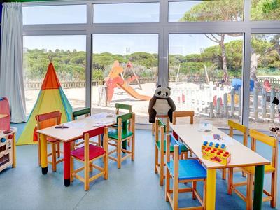 Heller Kinderspielraum mit bunten Tischen und Stühlen, direkter Blick auf den Spielplatz im Freien.