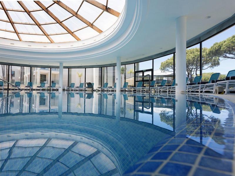 Innenaufnahme eines runden Hallenbads mit Glasdach und großen Fenstern, umgeben von Liegestühlen.