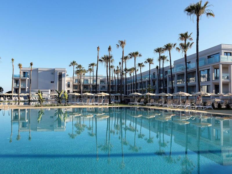 Großes Hotel mit Pool, umgeben von Palmen und Sonnenliegen bei klarem Himmel.