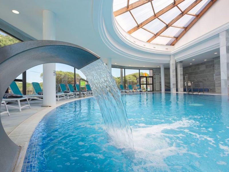 Innenpool mit Wasserfall, Liegestühlen und großen Fenstern unter einem Glasdach.