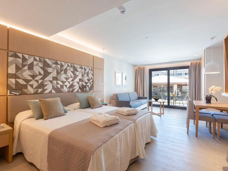 Helles Hotelzimmer mit zwei Betten, Sofa, Schreibtisch und Balkon mit Blick nach draußen.