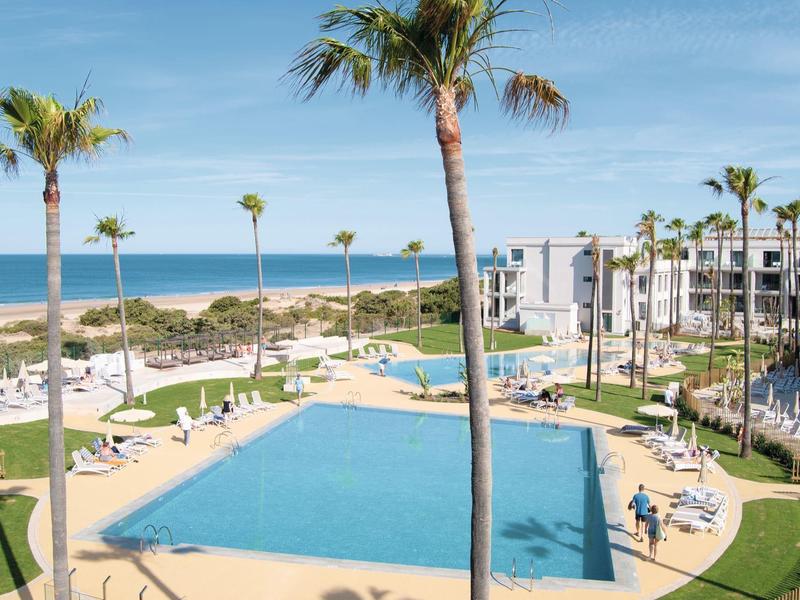 Hotelanlage mit großem Pool, Palmen und Meerblick bei klarem Himmel.