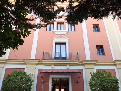 Mehrstöckiges Gebäude mit rosafarbenem Anstrich, großen Fenstern und Treppe, eingerahmt von Bäumen.