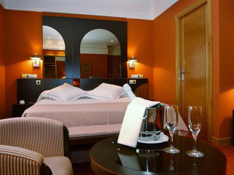 Gemütliches Hotelzimmer mit Doppelbett, orangen Wänden, Sessel und Tisch mit Champagner und Gläsern.