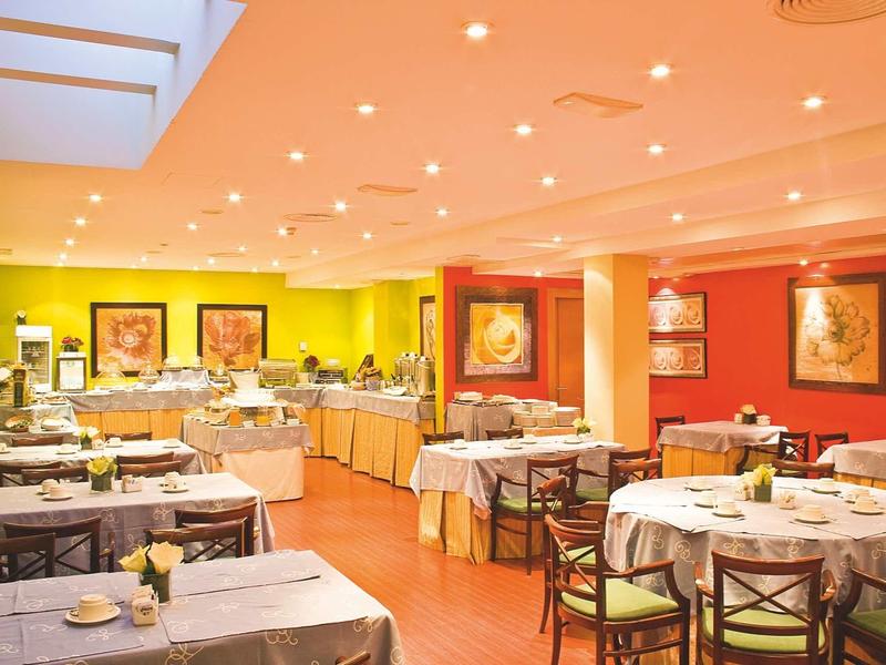 Hell beleuchteter Speisesaal mit gedeckten Tischen, Stühlen und einem Buffetbereich im Hintergrund.