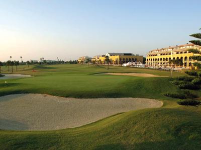 Grüner Golfplatz mit Bunker, Palmen und mehrstöckigen Gebäuden im Hintergrund bei klarem Himmel.