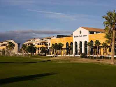 Großes Hotelgebäude mit gelber Fassade und Palmen vor grünem Rasen unter blauem Himmel.