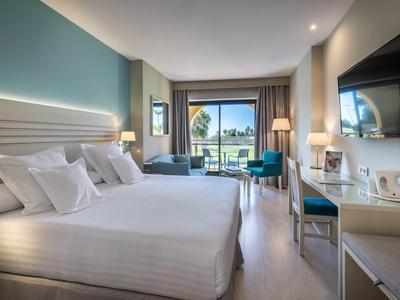 Modernes Hotelzimmer mit großem Bett, zwei Stühlen, Schreibtisch, Flatscreen-TV und Balkon mit Palmenblick.