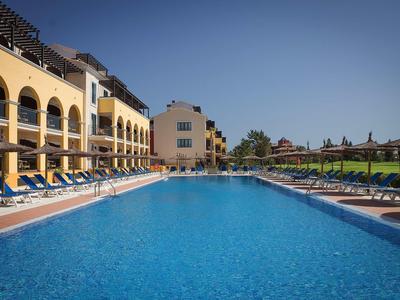 Großer blauer Pool mit Liegestühlen und gelben Gebäuden unter klarem blauem Himmel.