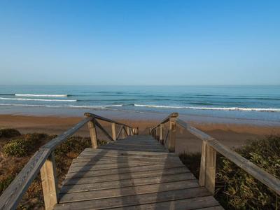 Holzsteg führt zu einem Strand mit Wellen, umgeben von Sanddünen und blauem Himmel.