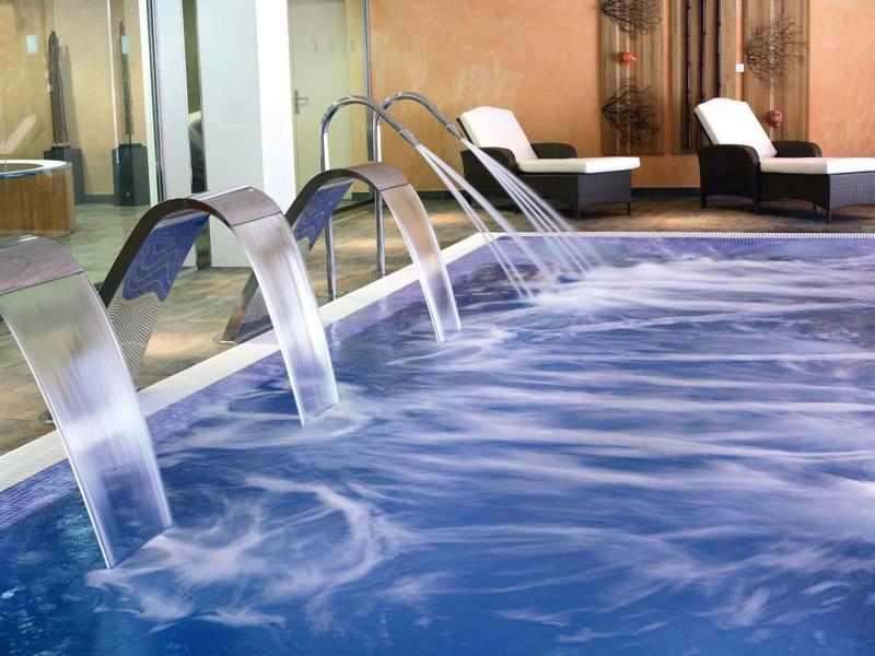 Moderner Pool mit drei Wasserspeiern und Liegen neben glasener Wand in warmem Ambiente.