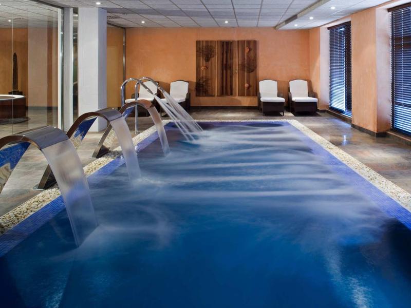 Innenpool mit Edelstahl-Wasserfontänen, blauer Wasseroberfläche, Holzpaneelen und weißen Liegestühlen.