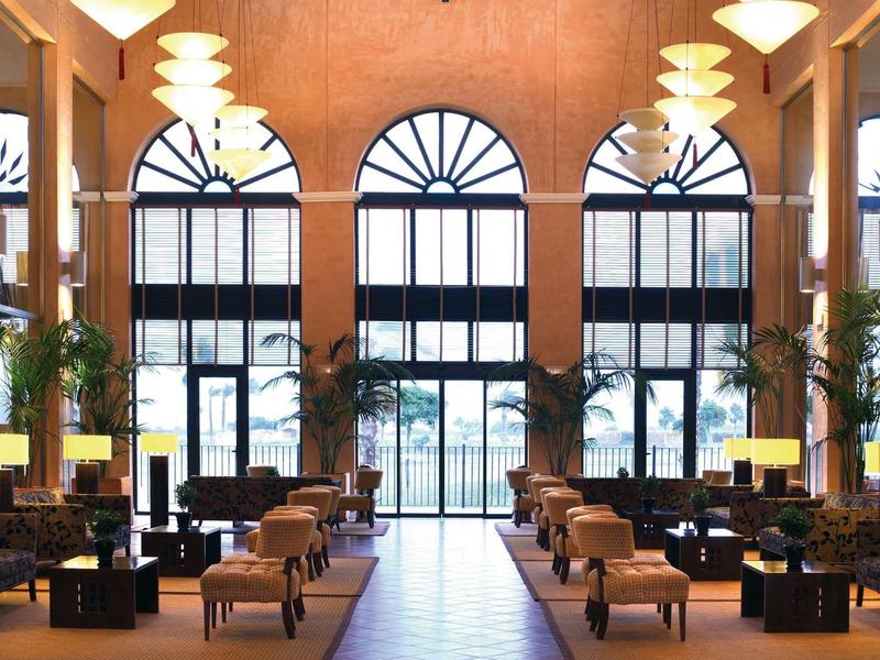 Großer heller Raum mit hohen Rundfenstern, gemütlichen Sitzgruppen und Pflanzen an den Wänden.