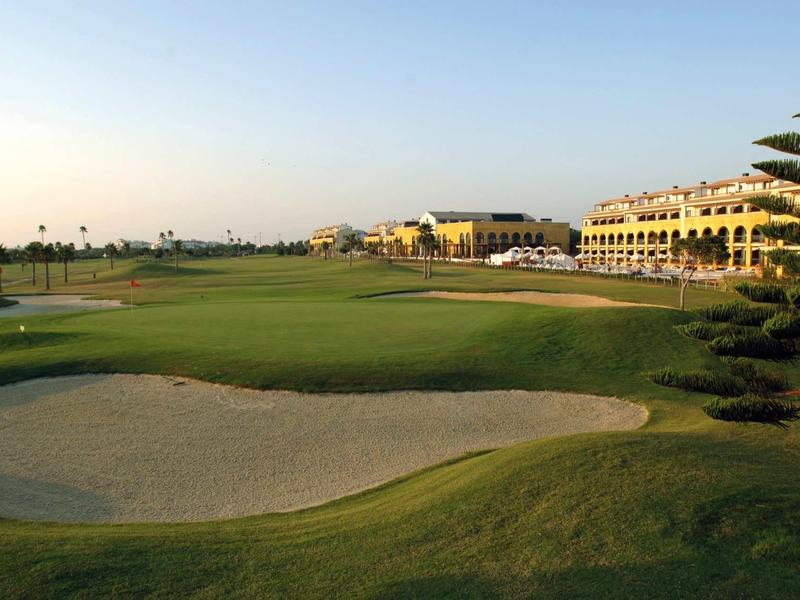 Grüner Golfplatz mit Bunker, Palmen und mehrstöckigen Gebäuden im Hintergrund bei klarem Himmel.