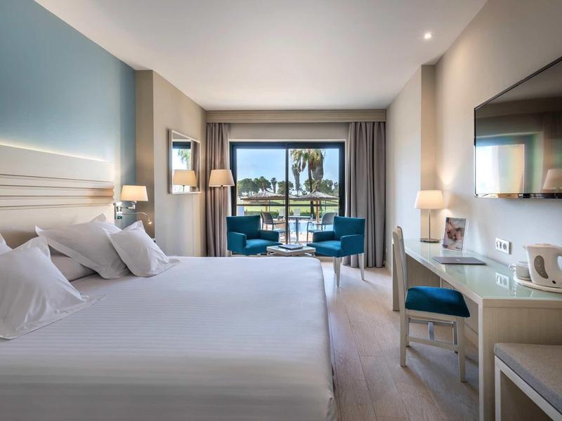 Helles Doppelzimmer mit weißer Bettwäsche, blauen Stühlen und Meerblick durch große Fenster.