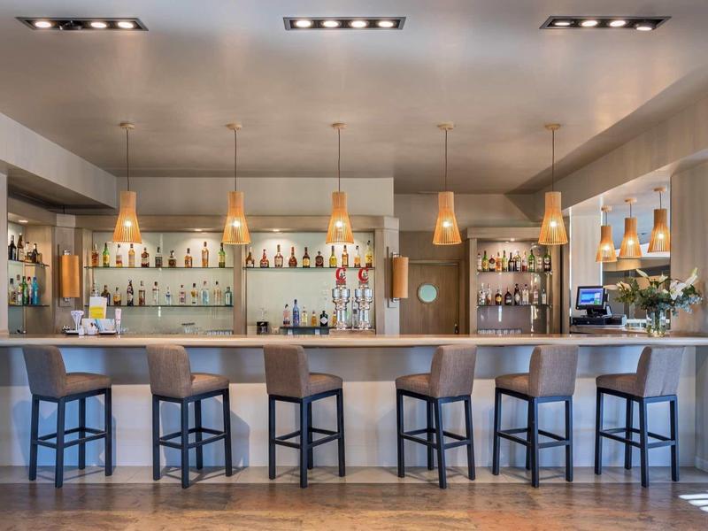 Moderner Barbereich mit fünf hohen Stühlen, Holztheke, flachen Pendelleuchten und gut bestücktem Regal.