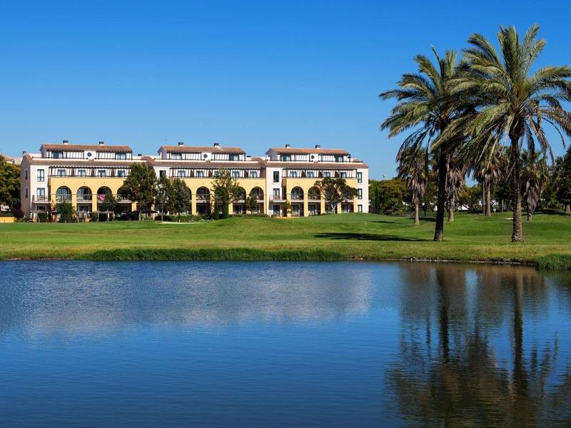 Großes gelbes Gebäude mit Säulen, Palmen und Teich unter klarem blauem Himmel.