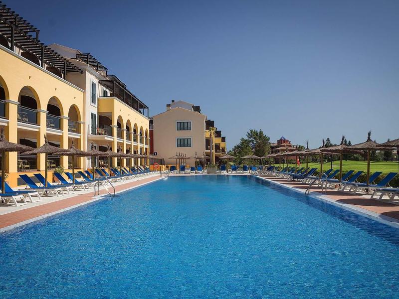 Großer blauer Pool mit Liegestühlen und gelben Gebäuden unter klarem blauem Himmel.