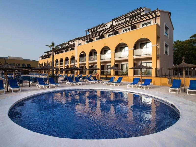Rundes Schwimmbecken vor gelbem Hotel mit blauen Liegestühlen und klarem Himmel im Hintergrund.