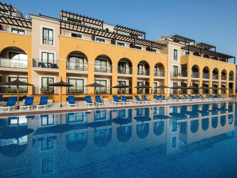 Großes gelbes Hotelgebäude mit blauen Liegen neben einem klaren Schwimmbecken unter blauem Himmel.