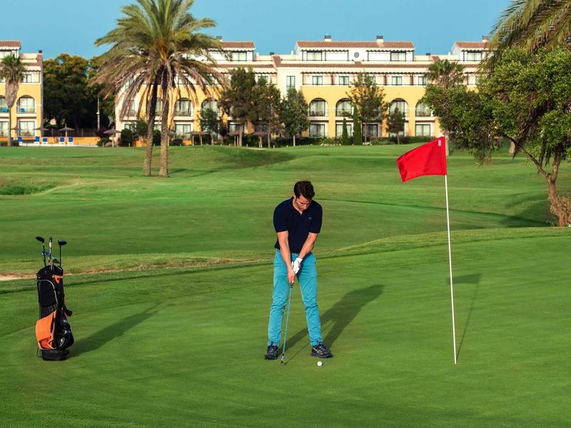 Mann spielt Golf auf grünem Rasen mit rotem Fahnenstock und Palmen im sonnigen Hintergrund.