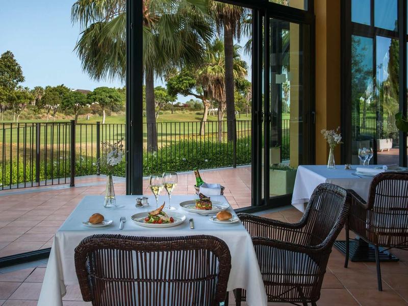 Gemütliches Restaurant mit Tisch am Fenster, Blick auf Terrasse, Palmen und Grünfläche draußen.