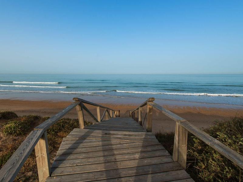 Holzsteg führt zu einem Strand mit Wellen, umgeben von Sanddünen und blauem Himmel.