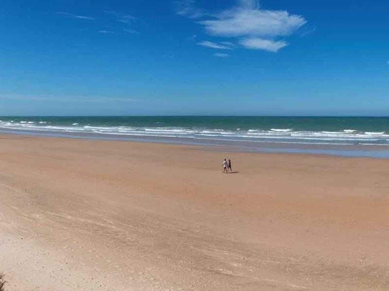 Breiter Sandstrand mit sanften Wellen, blauem Himmel und wenigen Wolken am Meer.