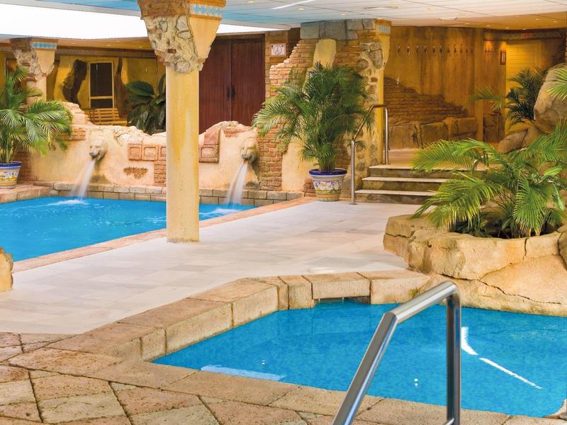 Innen-Schwimmbad mit hellem Steinmosaik, zwei Becken, Palmen und warmem Licht.