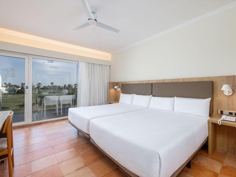 Helles Hotelzimmer mit zwei Betten, großem Balkon, Holzboden und Deckenventilator.