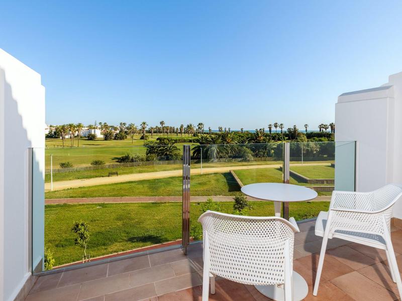Balkon mit zwei weißen Stühlen und Tisch, Blick auf grünen Golfplatz und blauen Himmel.
