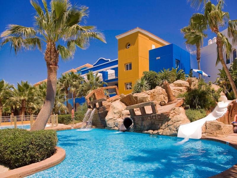 Ein blauer Pool mit Wasserrutsche und Felsen, umgeben von Palmen und bunten Gebäuden.