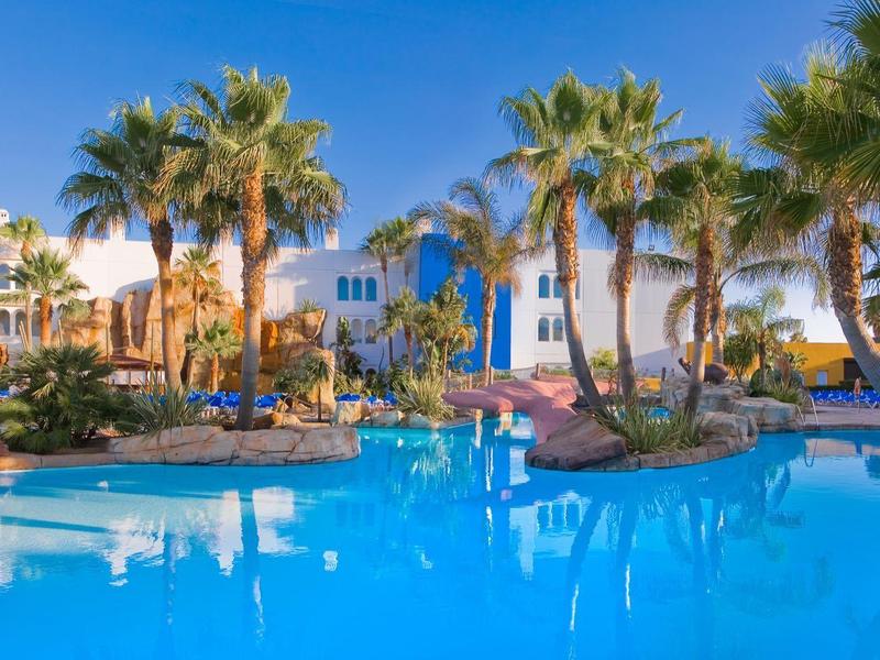 Blauer Pool mit Palmen und weißen Gebäuden unter klarem Himmel an sonnigem Tag.