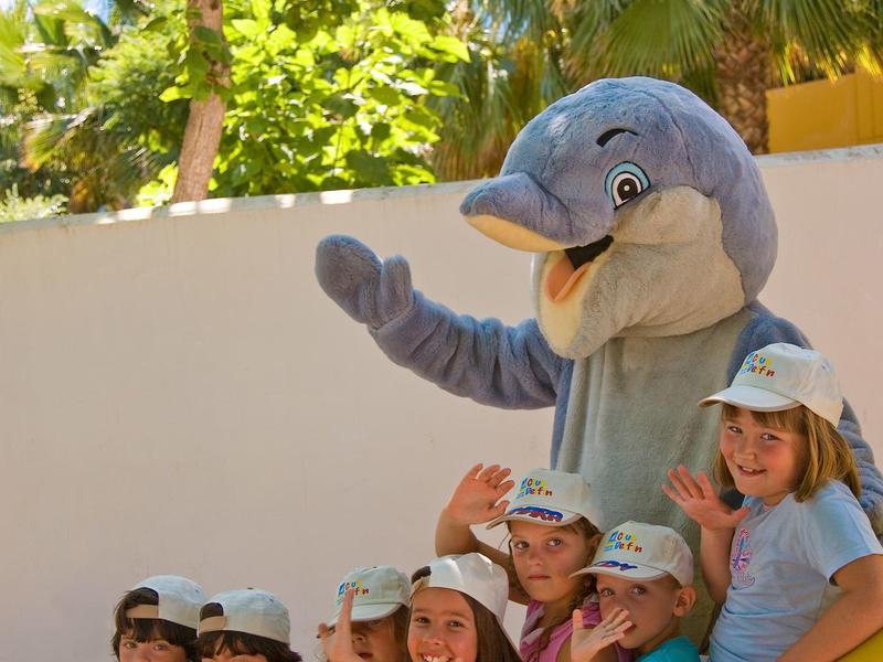 Kinder sitzen fröhlich auf einer gelben Rutsche, neben einer Person im Delfin-Kostüm.