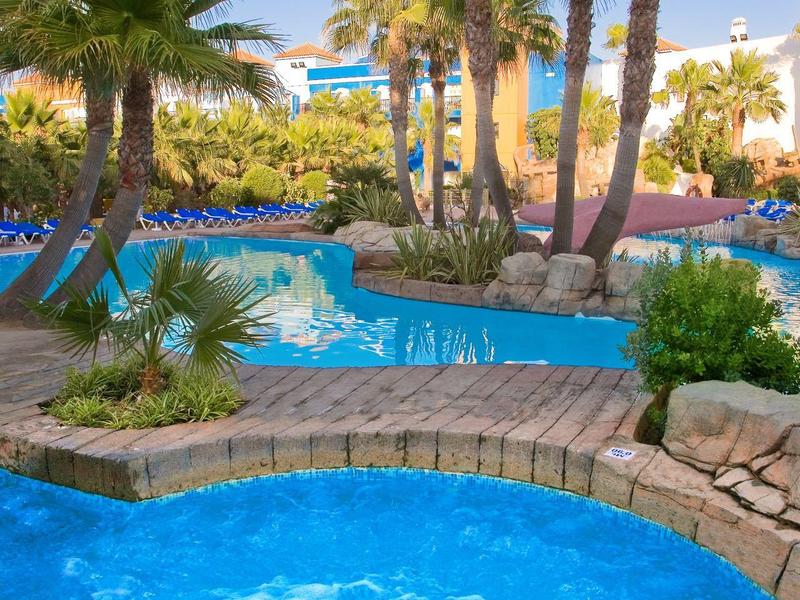 Blauer Pool mit klarem Wasser, umgeben von Palmen und grünen Pflanzen, bei sonnigem Himmel.