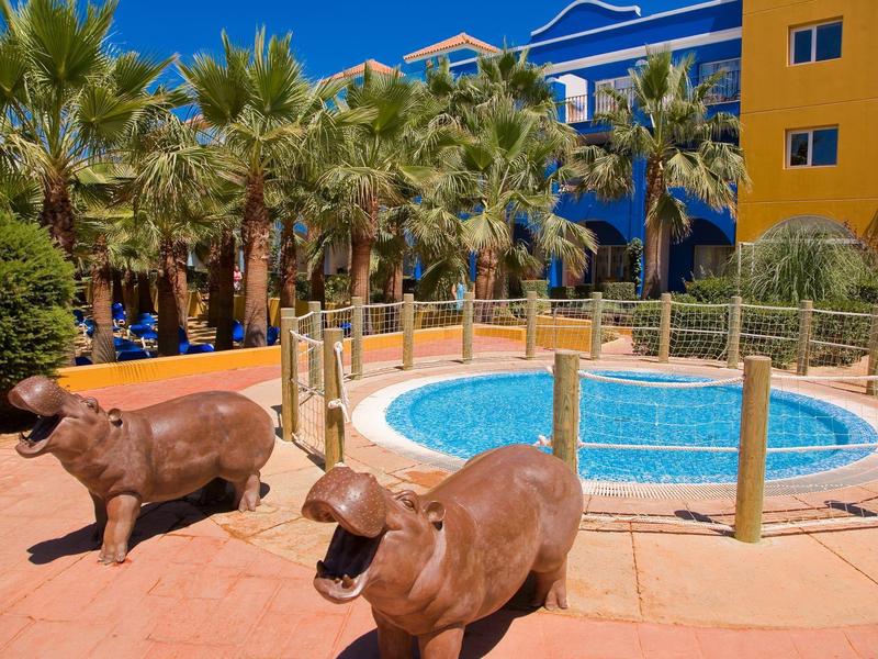 Poolbereich mit Palmen, blauen und gelben Gebäuden, und zwei Flusspferd-Skulpturen aus Bronze.