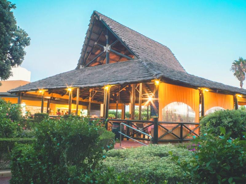 Ein großes, gelb beleuchtetes Haus mit braunem Dach, umgeben von grünen Pflanzen und Bäumen.