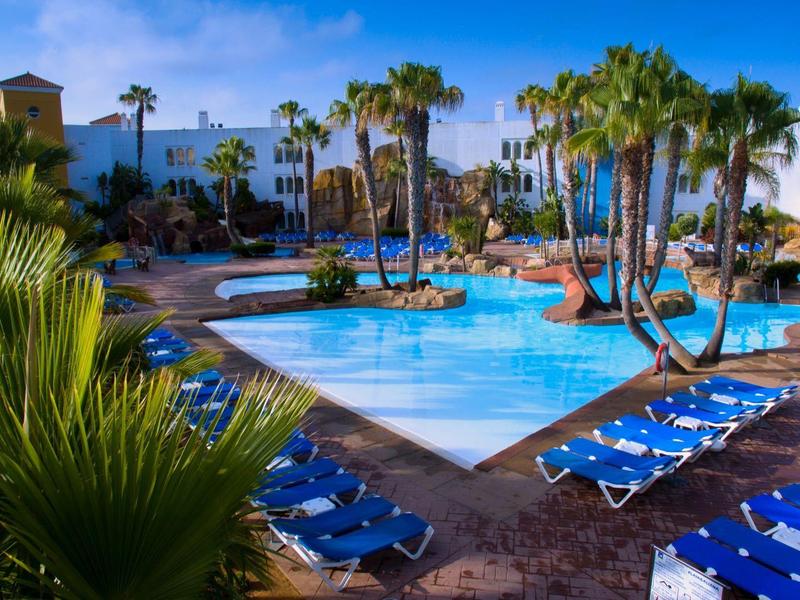 Großer Pool mit blauer Wasserfläche, umgeben von Palmen, blauen Liegestühlen und weißen Gebäuden.