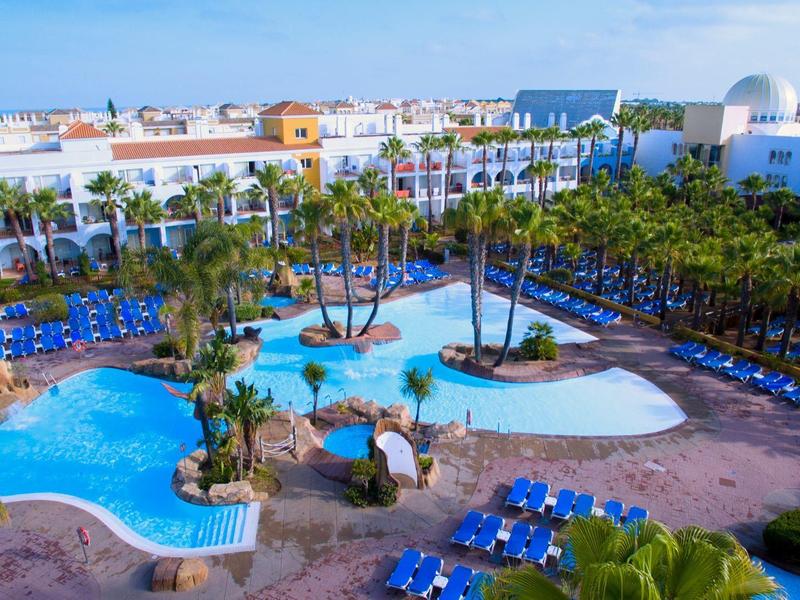 Großer Hotelpool mit Palmen, blauen Liegestühlen und weißem Hotelgebäude im sonnigen Wetter.