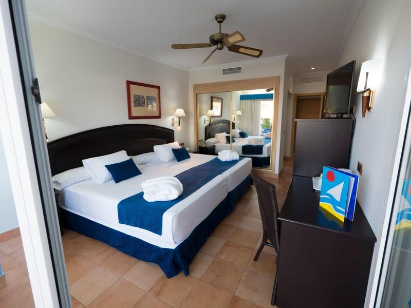 Großes, helles Hotelzimmer mit Doppelbett, Schreibtisch, Deckenventilator und Blick auf Wohnbereich.