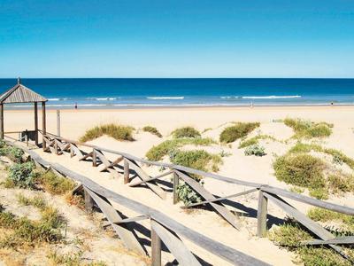 Strand mit hellem Sand, blauen Wasser, Dünen und Holzsteg mit Geländer und kleiner Überdachung.