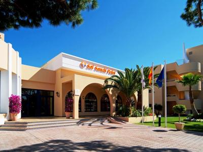 Eingang eines Gebäudes mit Bögen, Palmen und Flaggen vor klarem blauem Himmel.