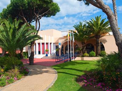 Ein sonniger Hotelgarten mit Palmen, grünem Rasen, bunten Blumen und einem mediterranen Gebäude.