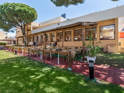 Außenansicht eines Hotels mit Terrasse, Tischen und Stühlen unter Bäumen bei Sonnenschein.