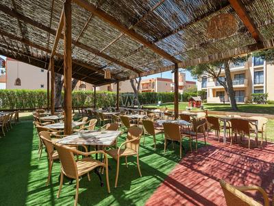 Überdachte Terrasse mit Tischen und Stühlen in Hotelanlage mit Rasenfläche und Gebäuden im Hintergrund.