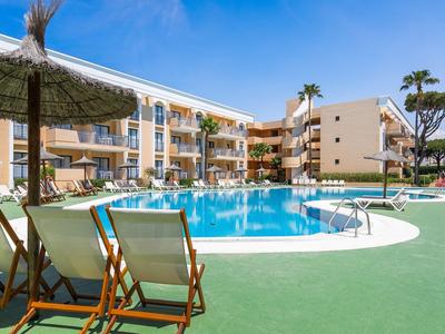 Hotel mit Außenpool, Liegestühlen und Sonnenschirmen bei blauem Himmel.