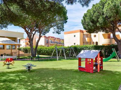 Kinderspielplatz mit Rutschbahn, Schaukel, Wippe und einem kleinen roten Spielhaus auf grünem Gras.